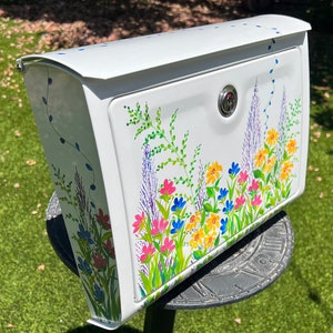 Op de afbeelding: Witte metalen brievenbus met een handgeschilderd bloemenontwerp. De brievenbus heeft een kleurrijk ontwerp van wilde bloemen in roze, geel, blauw en paars, met groene bladeren. De brievenbus heeft een slot en is gemonteerd op een zwarte metalen standaard.