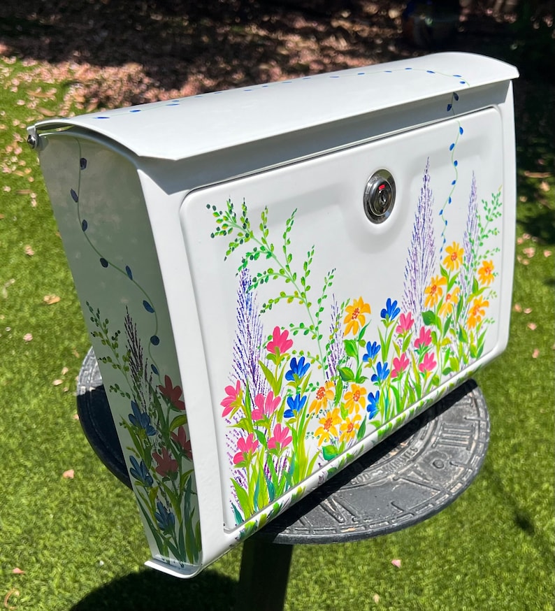 Op de afbeelding: Witte metalen brievenbus met een handgeschilderd bloemenontwerp. De brievenbus heeft een kleurrijk ontwerp van wilde bloemen in roze, geel, blauw en paars, met groene bladeren. De brievenbus heeft een slot en is gemonteerd op een zwarte metalen standaard.