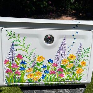 Op de afbeelding: Witte brievenbus met een bloemenontwerp. De brievenbus heeft een slot en is versierd met kleurrijke bloemen in roze, geel, blauw en paars, met groene bladeren. De bloemen zijn op de voorkant van de brievenbus geschilderd.