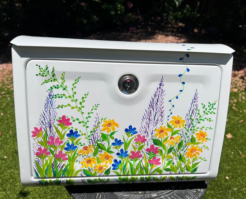 Op de afbeelding: Witte brievenbus met een bloemenontwerp. De brievenbus heeft een slot en is versierd met kleurrijke bloemen in roze, geel, blauw en paars, met groene bladeren. De bloemen zijn op de voorkant van de brievenbus geschilderd.
