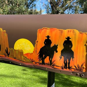 Puede incluir: Un buzón marrón con una escena del desierto pintada. La obra de arte representa una puesta de sol con dos vaqueros a caballo. El fondo presenta un sol amarillo, formaciones rocosas naranjas y amarillas y cactus. El buzón está montado en un poste de metal.