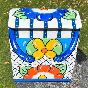 May include: A white mailbox with a blue, yellow, orange, and green floral design. The mailbox has a blue trim and a white background with a blue and white checkered pattern.