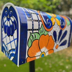 May include: A hand-painted mailbox with a vibrant floral design. The mailbox features a white base with blue and orange flowers, green leaves, and blue accents. The design is on a curved metal surface.