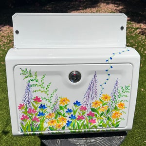 Op de afbeelding: Witte brievenbus met een bloemenontwerp. De brievenbus heeft een slot en een scharnierende deur. Het bloemenontwerp omvat roze, gele en blauwe bloemen met groene bladeren. Een wijnstok met blauwe accenten loopt langs de zijkant.