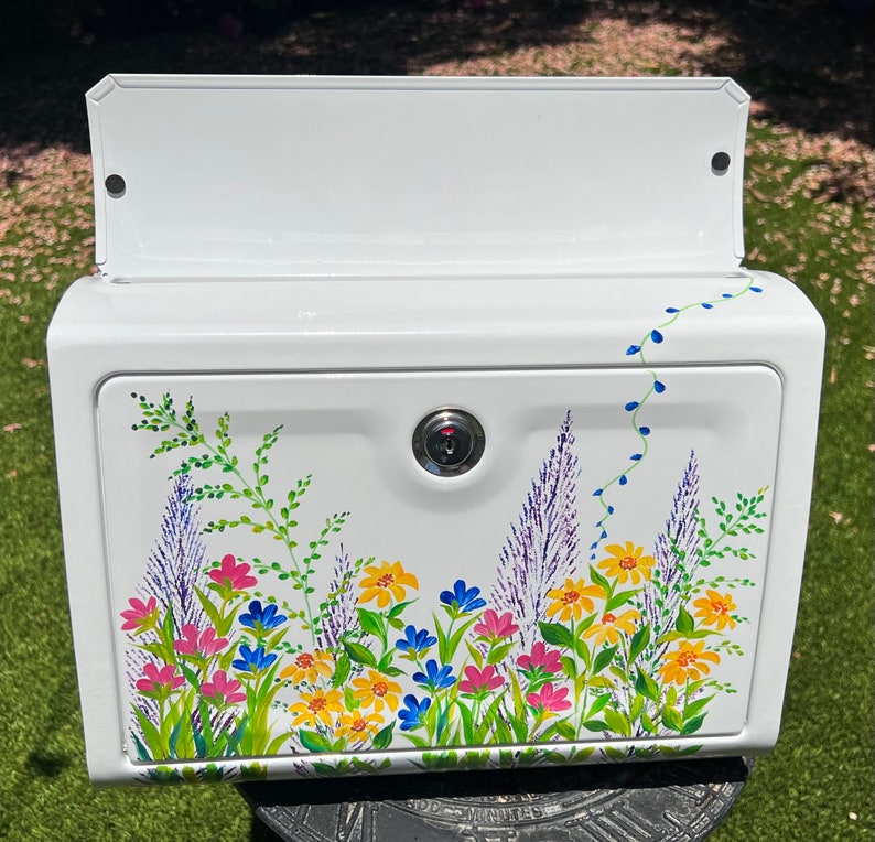 Op de afbeelding: Witte brievenbus met een bloemenontwerp. De brievenbus heeft een slot en een scharnierende deur. Het bloemenontwerp omvat roze, gele en blauwe bloemen met groene bladeren. Een wijnstok met blauwe accenten loopt langs de zijkant.
