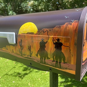 Puede incluir: Un buzón de metal marrón con una escena desértica pintada. La obra de arte representa un sol amarillo, cactus y dos vaqueros a caballo con lazos. El buzón tiene un asa blanca.