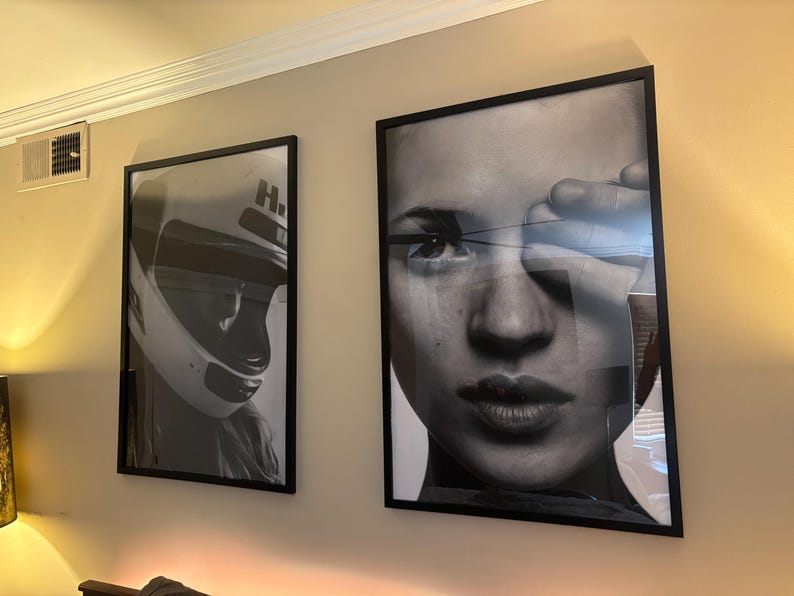 May include: Two framed black and white art prints hanging on a beige wall. One print features a racing helmet, and the other is a close-up portrait of a person's face. Both prints are in black frames.