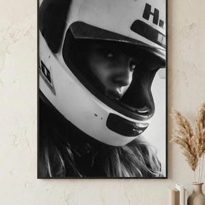 May include: Black and white framed print of a person wearing a white motorcycle helmet. The helmet has a dark visor and the letter "H" on the side. The person's face is partially visible, with long hair flowing out from under the helmet.