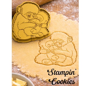 May include: A yellow cookie cutter in the shape of two hugging monkeys, next to a cookie cut-out of the same design. The text "Stampin' Cookies" is in black script. The scene is set on a wooden surface with a rolling pin.