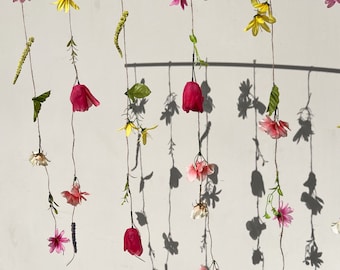 May include: A white wall with strings of pink, yellow, and red artificial flowers hanging from a wooden rod. The flowers are arranged in a cascading pattern, creating a whimsical and colorful display.