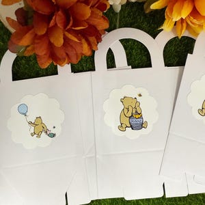 May include: White gift bags with arched handles and Winnie the Pooh character stickers. One sticker shows Pooh with a blue balloon, and another shows Pooh with a honey pot. The bags are on a green surface with orange and yellow flowers.