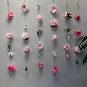 May include: A wall decoration with multiple strands of pink flowers and leaves hanging from a wooden dowel.
