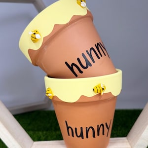 May include: Two terracotta flower pots stacked. The pots have a yellow rim with a wavy edge and small bee decorations. The word "hunny" is written in black on the front of each pot. The pots are on a wooden shelf with a honeycomb design.