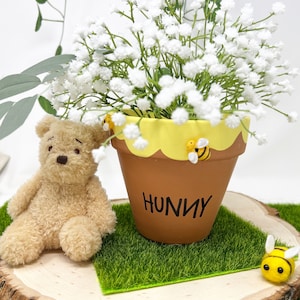 May include: A brown terracotta flower pot with a yellow rim and a white background. The pot is decorated with three small yellow bees and the word "Hunny" written in black. The pot is filled with white baby's breath flowers and is sitting on a wooden slice with green grass.