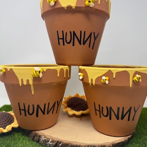 May include: Three terracotta flower pots with yellow honey-like paint dripping down the sides. Each pot has the word "Hunny" written in black paint and has small yellow and black bee figurines attached.