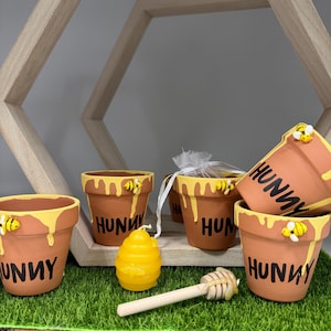 May include: Terracotta flower pots with yellow honey-like paint dripping down the sides, each with the word "HUNNY" and a bee. A yellow honeycomb candle and a wooden honey dipper are also present. The items are arranged on a bed of green artificial grass.