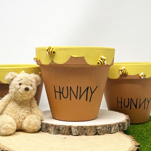 May include: Three terracotta flower pots painted with a yellow honey design and the word "Hunny" in black. Each pot has small yellow and black bee figurines attached to the rim.