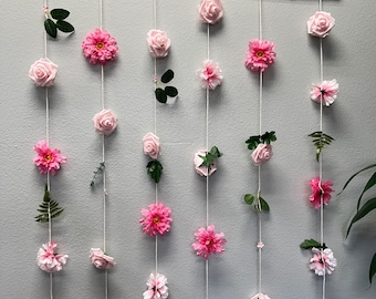 May include: A pink flower garland with a wooden dowel at the top. The garland is made of pink flowers and greenery, and is hanging on a white wall.