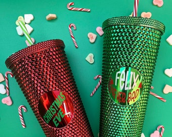May include: Two studded tumblers, one red and one green, with straws, surrounded by candy hearts and canes on a green background. The red tumbler has a sticker that says "Christmas Eve". The green tumbler has a sticker that says "Felix Never Bad".