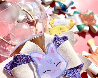 Pode incluir: Um laço branco e roxo com glitter e um amuleto de plástico transparente de um gato azul claro com um rosto sorridente. O amuleto do gato tem um contorno branco e olhos pretos.