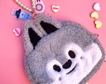 May include: A gray plush wolf-shaped coin purse with black ears and a white face, featuring a pearl beaded strap and a keychain with the text "STAY". Pink and white heart-shaped candies with "XOXO" text are scattered around the purse.