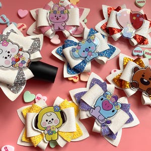 May include: A selection of handmade hair bows in various colours, with glitter accents and cartoon character embellishments. Each bow has a unique design with a central character, surrounded by decorative elements. The bows are arranged on a pink surface.