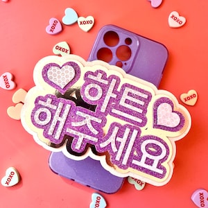 May include: A purple smartphone case featuring a large, layered, glittery purple and white decorative topper with Korean text that reads "하트 해주세요" and heart icons, surrounded by small, scattered, heart-shaped candies labeled "XOXO".