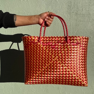May include: A red and gold woven tote bag with two handles. The bag is made of a woven plastic material and has a geometric pattern.