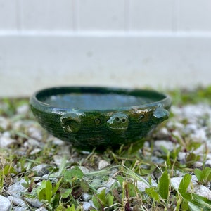 Op de afbeelding: Een handgemaakte, groene keramische kom met een varkensgezichtontwerp. De kom heeft een ronde vorm met een getextureerde buitenkant en een glanzend, donkergroen glazuur. De snuit en oren van het varken zijn duidelijk zichtbaar aan de voorkant van de kom.