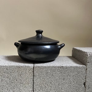 Może przedstawiać: Czarny ceramiczny garnek do gotowania z pokrywką, z dwoma małymi uchwytami po bokach. Garnek stoi na szarym bloku betonowym, z neutralnym tłem. Garnek ma gładkie, błyszczące wykończenie.
