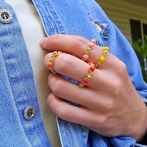 May include: Three gold rings with colorful beads. The rings are stacked on a finger. The beads are pink, red, yellow, and green.