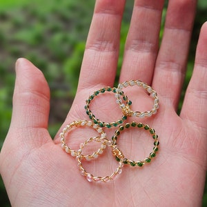 Puede incluir: Cinco anillos de oro con cuentas de diferentes colores. Los anillos están hechos con pequeñas cuentas redondas en tonos blancos y verdes. Los anillos están apilados uno encima del otro en una mano.