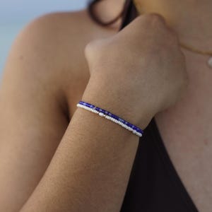 May include: A beaded bracelet with a pattern of white and blue beads. The bracelet is worn on a person's wrist. The background is blurred, suggesting an outdoor setting. The bracelet is a piece of jewelry.