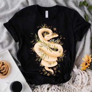 May include: Black t-shirt with a white snake wrapped around a bouquet of yellow and white flowers. The snake is surrounded by green leaves and the flowers have yellow centers.
