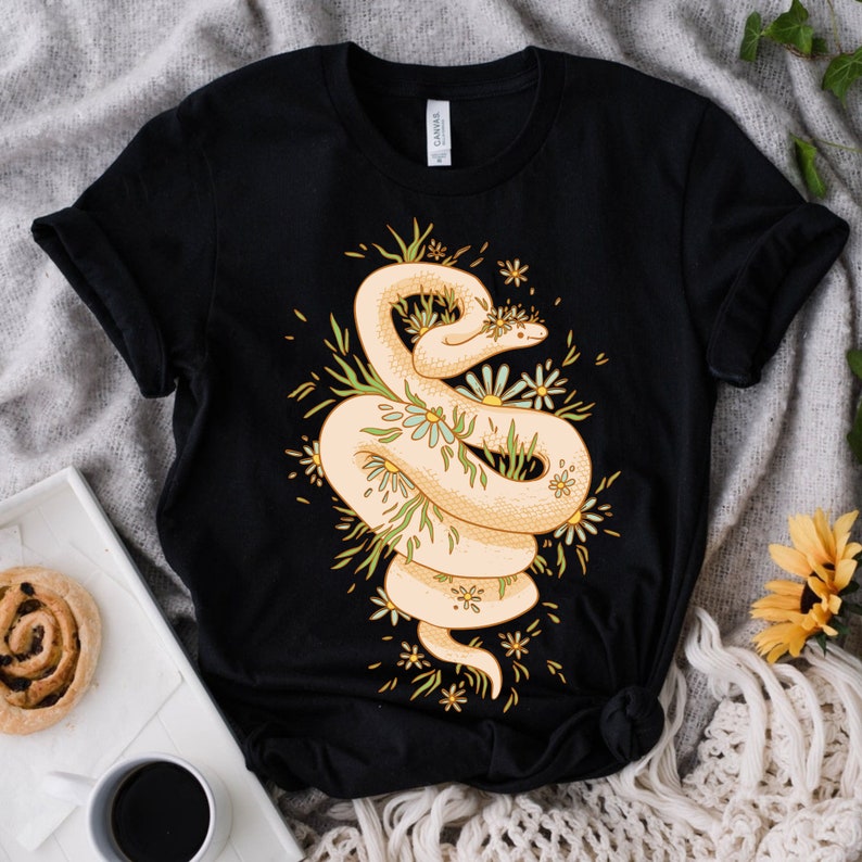 May include: Black t-shirt with a white snake wrapped around a bouquet of yellow and white flowers. The snake is surrounded by green leaves and the flowers have yellow centers.