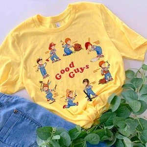 May include: Yellow t-shirt with a graphic of various characters from the movie Child's Play. The characters are all depicted as "good guys" and have different professions. The text "Good Guys" is printed in red on the shirt.