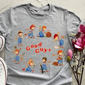 May include: Gray t-shirt with a graphic print of eight cartoon characters in different outfits and poses. The characters are all holding tools or playing sports. The text "Good Guys" is printed in large letters below the characters.