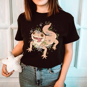 May include: A black t-shirt with a cartoon illustration of a lizard wearing a flower crown. The lizard is brown with a pink tongue and is surrounded by flowers.