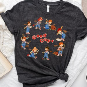 May include: A dark gray t-shirt with a graphic print of various cartoon characters holding tools and weapons. The text "Good Guys" is printed in red on the shirt.