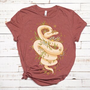 May include: A maroon t-shirt with a white snake design. The snake is curled up in a circle and surrounded by green leaves and white flowers.