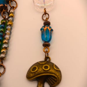 Może przedstawiać: Wielorzędowy naszyjnik z niebieskimi i zielonymi koralikami, przezroczystą, tęczową koralikiem i złotym zawieszką w kształcie grzyba.