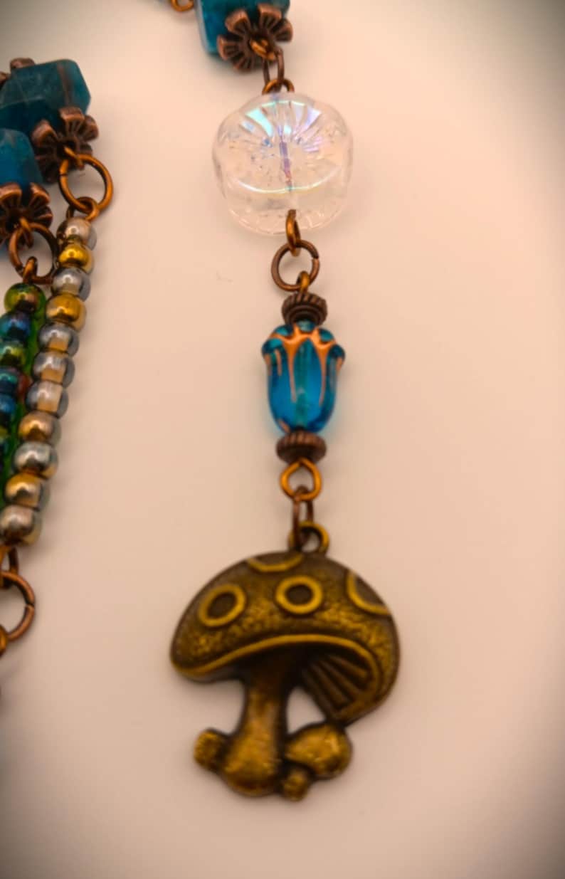 Może przedstawiać: Wielorzędowy naszyjnik z niebieskimi i zielonymi koralikami, przezroczystą, tęczową koralikiem i złotym zawieszką w kształcie grzyba.