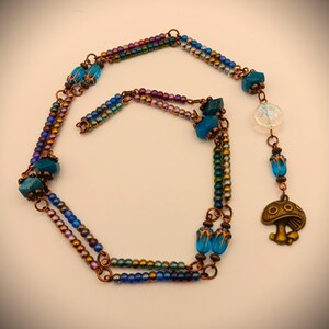 Może przedstawiać: Wielorzędowy naszyjnik z niebieskimi i tęczowymi koralikami, drutem miedzianym i zawieszką w kształcie grzyba z twarzą.