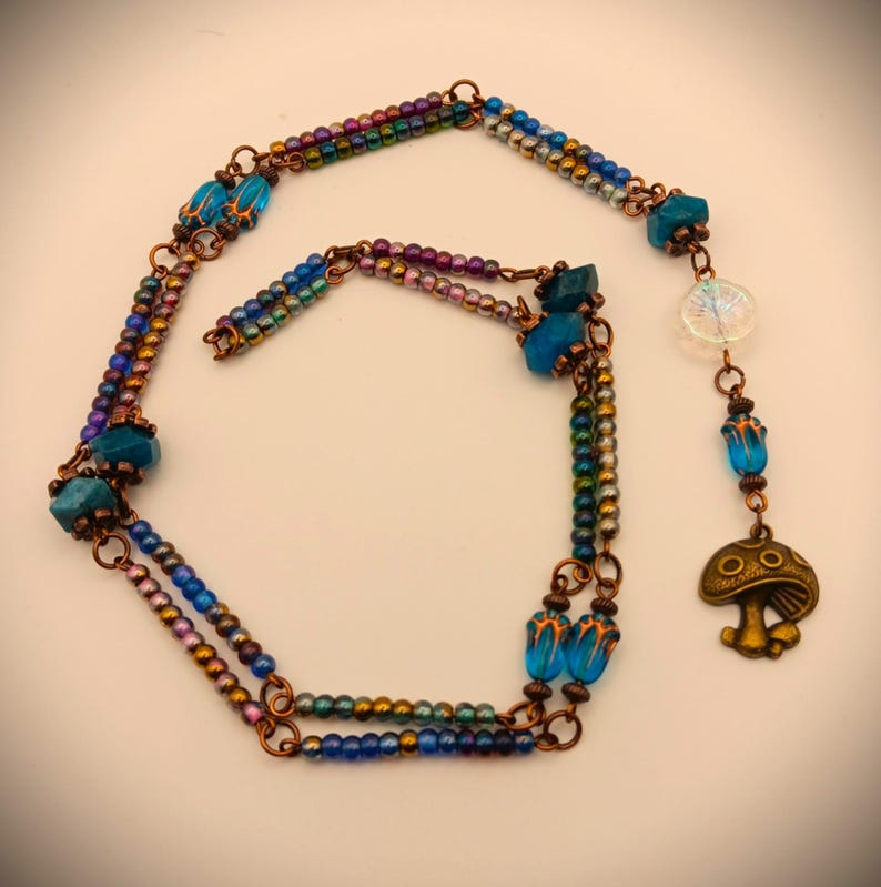 Może przedstawiać: Wielorzędowy naszyjnik z niebieskimi i tęczowymi koralikami, drutem miedzianym i zawieszką w kształcie grzyba z twarzą.