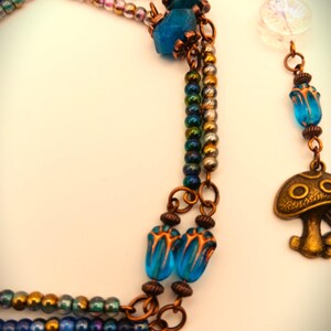 Może przedstawiać: Wielowarstwowy naszyjnik z miedzianym łańcuszkiem i niebieskimi szklanymi koralikami. Naszyjnik ma zawieszkę w kształcie grzyba i przezroczysty szklany koralik z wzorem płatka śniegu.