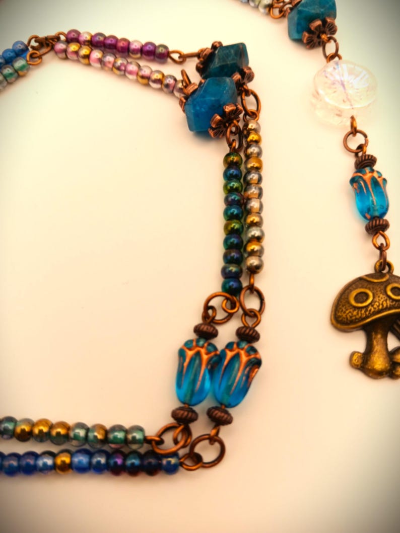 Może przedstawiać: Wielowarstwowy naszyjnik z miedzianym łańcuszkiem i niebieskimi szklanymi koralikami. Naszyjnik ma zawieszkę w kształcie grzyba i przezroczysty szklany koralik z wzorem płatka śniegu.