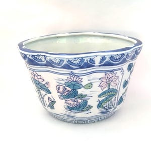 May include: A white ceramic planter with a blue rim and decorative floral and bird motifs. The planter features a scalloped edge and a detailed design of lotus flowers and ducks. The interior is white, and the exterior has a detailed pattern.