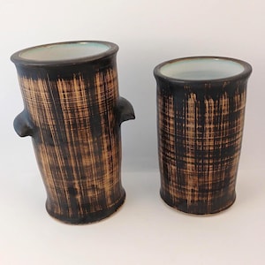 May include: Two cylindrical ceramic vases with a dark brown and tan cross-hatched brushstroke pattern. The taller vase features two small side handles, while both vessels have a smooth, light-coloured interior finish and a flared top rim.