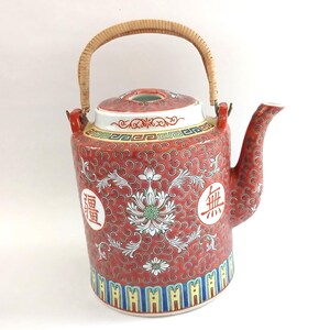 May include: Red ceramic teapot with a woven handle and lid. The teapot features floral and scroll patterns, with Chinese characters in white circles. The base has a colourful geometric border.
