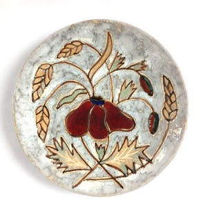 May include: A decorative ceramic plate with a floral design. The plate features a central red flower with a blue centre, surrounded by green leaves and tan stems. The plate has a light blue background with a speckled texture and a gold rim.